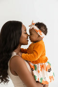 Maman et bébé fille orange BabyCoton — bébé debout soutenue, cardigan manches longues orange, robe carottes nœuds plissée et bandeau, fond blanc