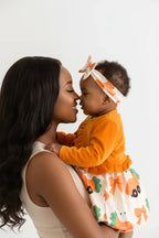 Maman et bébé fille orange BabyCoton — bébé debout soutenue, cardigan manches longues orange, robe carottes nœuds plissée et bandeau, fond blanc
