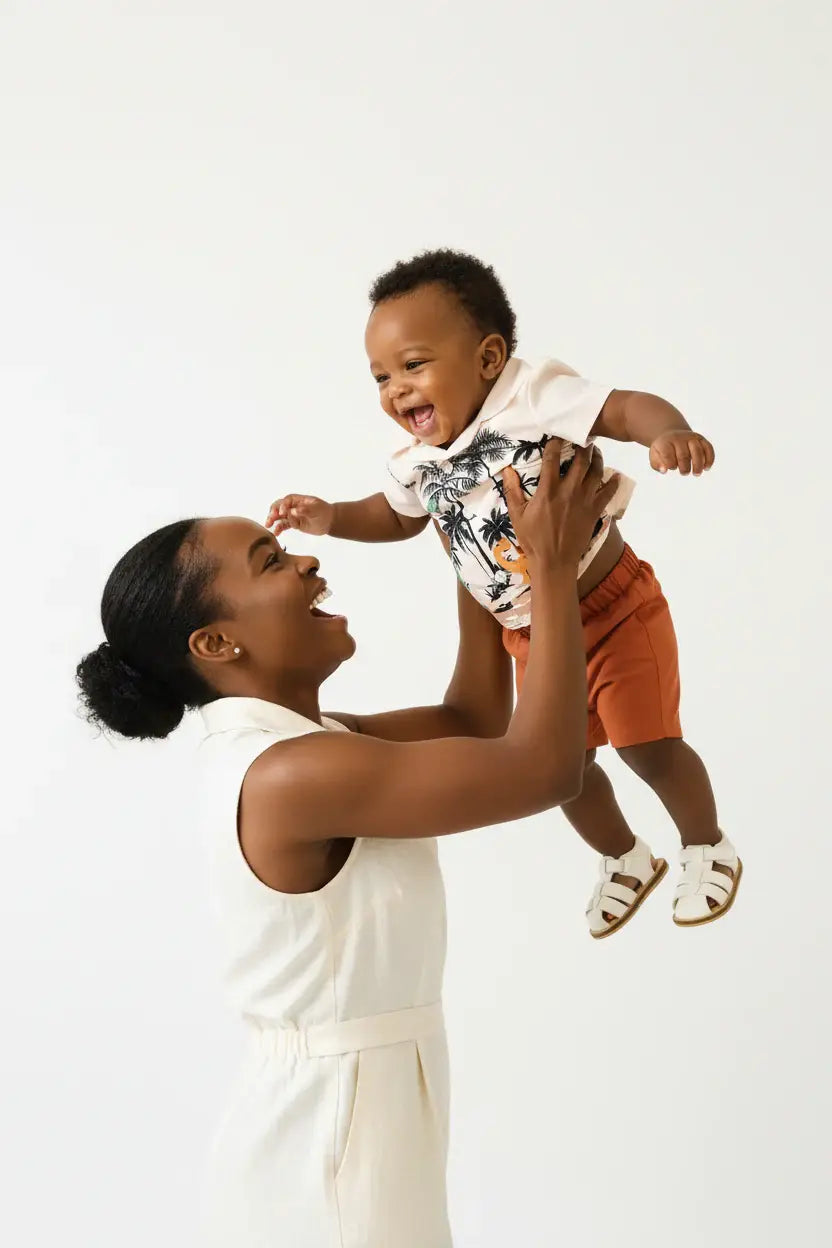 Maman soulevant bébé garçon en ensemble dinosaures palmiers BabyCoton — chemise polo beige dinos tropicaux et short terracotta, moment de joie fond blanc