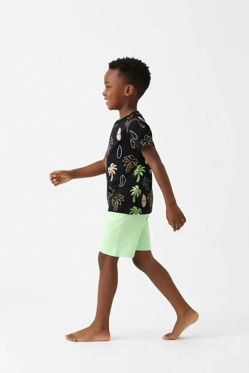 Vue de profil garçon en marche — ensemble palmiers tropicaux BabyCoton, t-shirt noir palmiers surf et short vert fluo, fond blanc