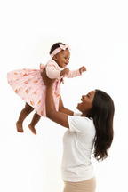 Maman soulevant bébé fille en ensemble rose BabyCoton — jupe plissée lapins carottes qui s'envole, cardigan volants et bandeau nœud, fond blanc