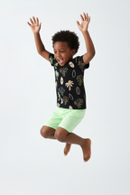 Garçon qui saute en ensemble palmiers tropicaux BabyCoton — t-shirt noir all-over palmiers surf et short vert fluo, mouvement dynamique fond blanc