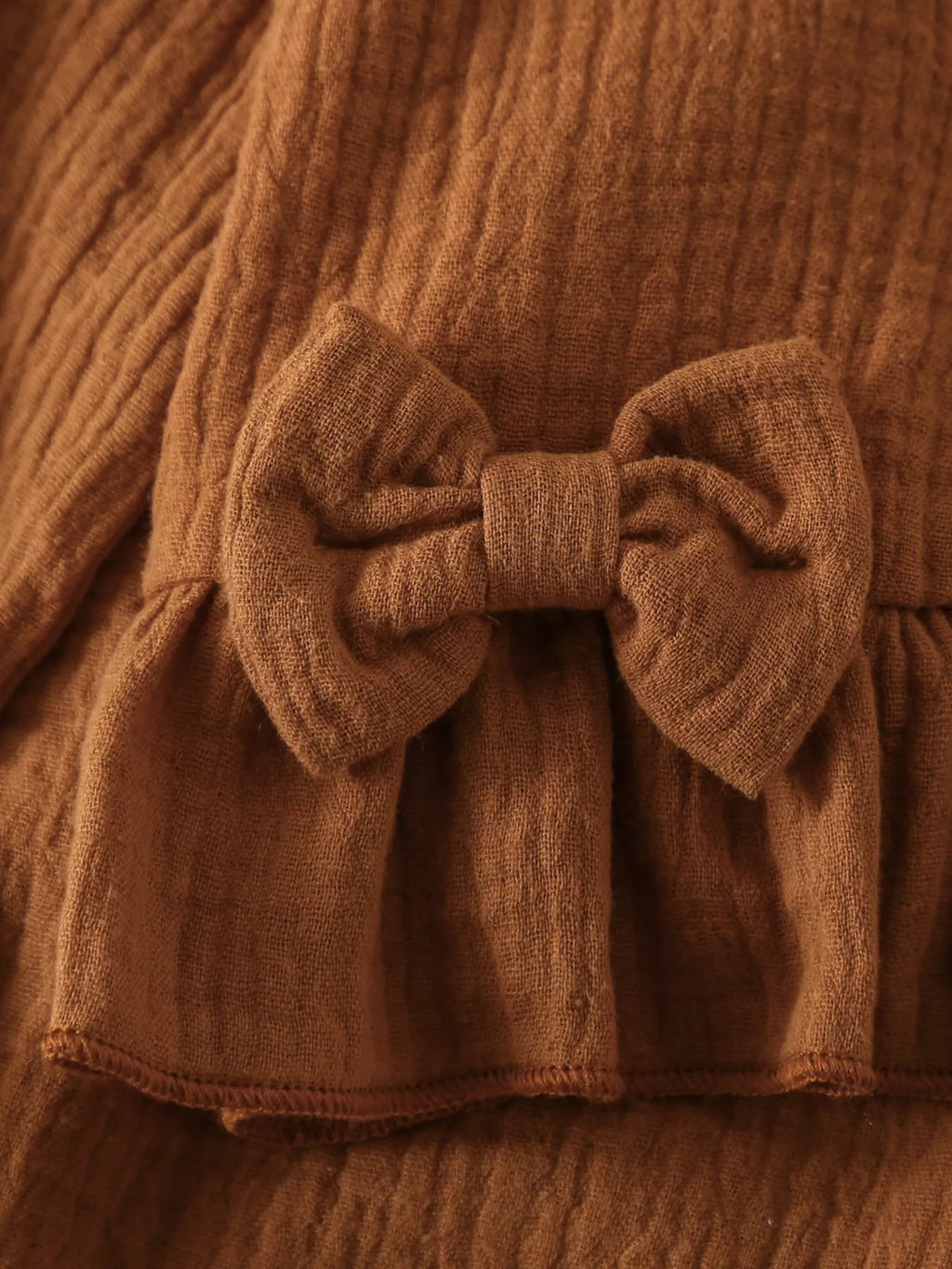 Détail rapproché des volants romantiques et du nœud décoratif rose poudré ornant l'ensemble bébé, mettant en valeur la finition soignée