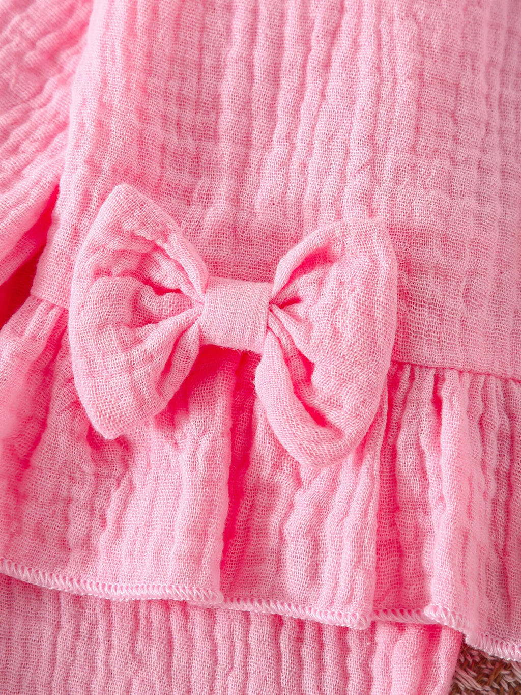 Détail rapproché des volants romantiques et du nœud décoratif rose poudré ornant l'ensemble bébé, mettant en valeur la finition soignée