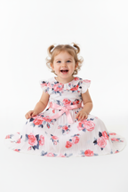 Fille en robe blanche plissée fleurie roses - col volant romantique ceinture satin rose - pose assise adorable expression douce - BabyCoton