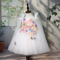 "Robe blanche de cérémonie pour fille avec appliqués faits main : cascade de fleurs 3D en tons pastel (rose, violet, jaune, bleu) ornées de perles et strass ✨ + papillons dorés brodés 🦋 - jupe en tulle vaporeux blanc, col à volants délicats, design féerique unique parfait pour baptême, mariage ou séance photo professionnelle, tailles 80-130cm chez BabyCoton Suisse 🌸"
