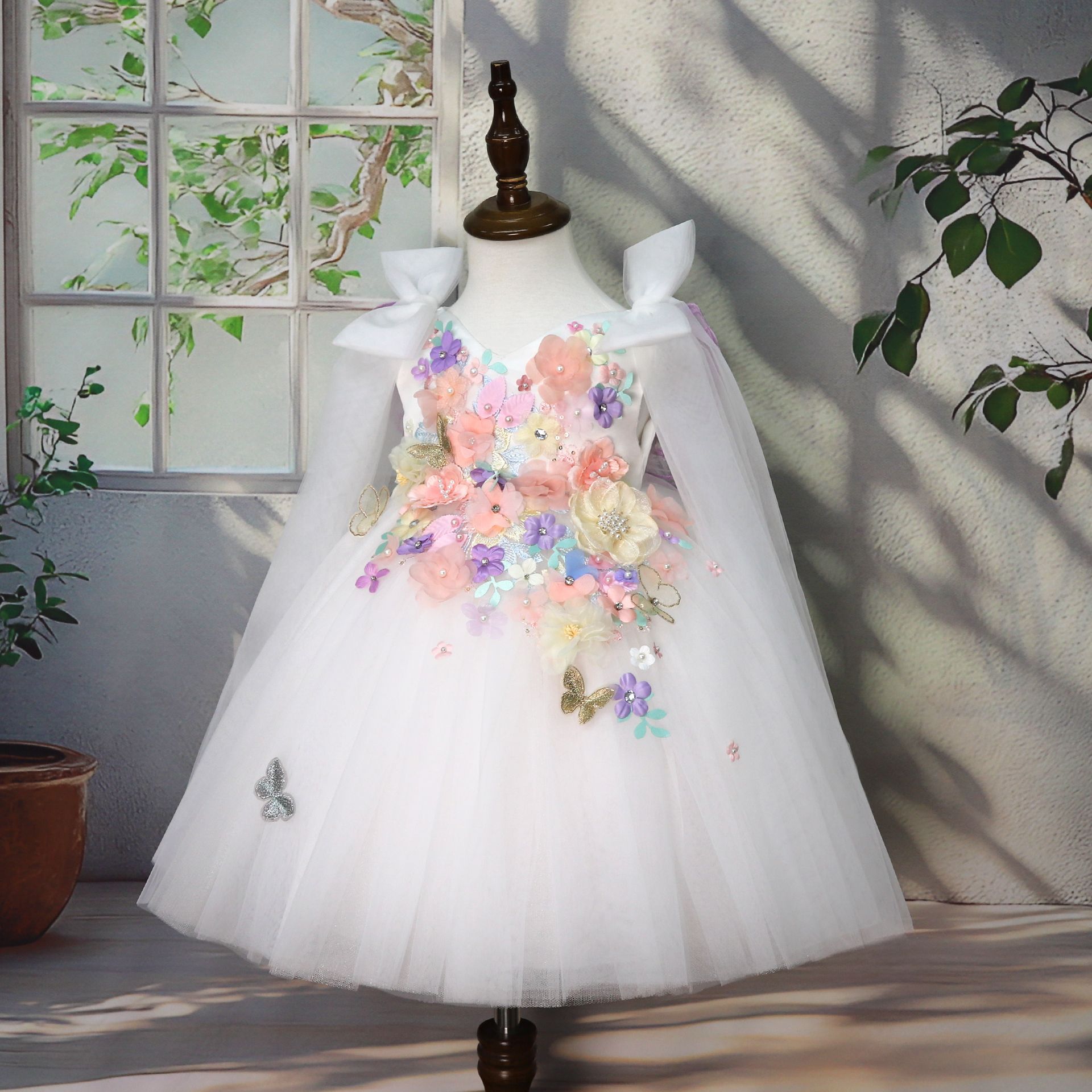 "Robe blanche de cérémonie pour fille avec appliqués faits main : cascade de fleurs 3D en tons pastel (rose, violet, jaune, bleu) ornées de perles et strass ✨ + papillons dorés brodés 🦋 - jupe en tulle vaporeux blanc, col à volants délicats, design féerique unique parfait pour baptême, mariage ou séance photo professionnelle, tailles 80-130cm chez BabyCoton Suisse 🌸"