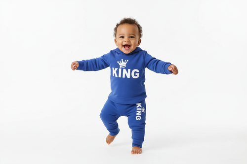 Bébé garçon d'origine diverse debout ou faisant ses premiers pas en portant un body hoodie bleu avec texte KING et couronne blanc et pantalon bleu assorti
