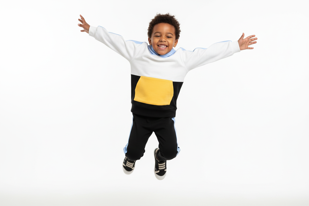 Enfant  sautant en l'air avec joie en portant un hoodie color block blanc, bleu ciel, jaune et noir avec pantalon noir à bandes bleues
