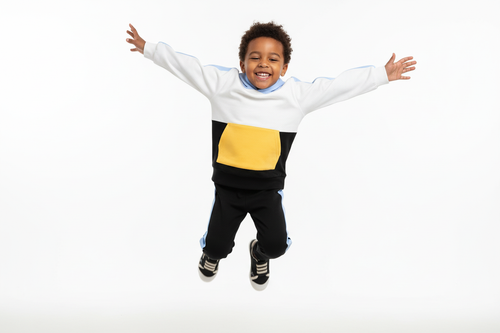 Enfant  sautant en l'air avec joie en portant un hoodie color block blanc, bleu ciel, jaune et noir avec pantalon noir à bandes bleues
