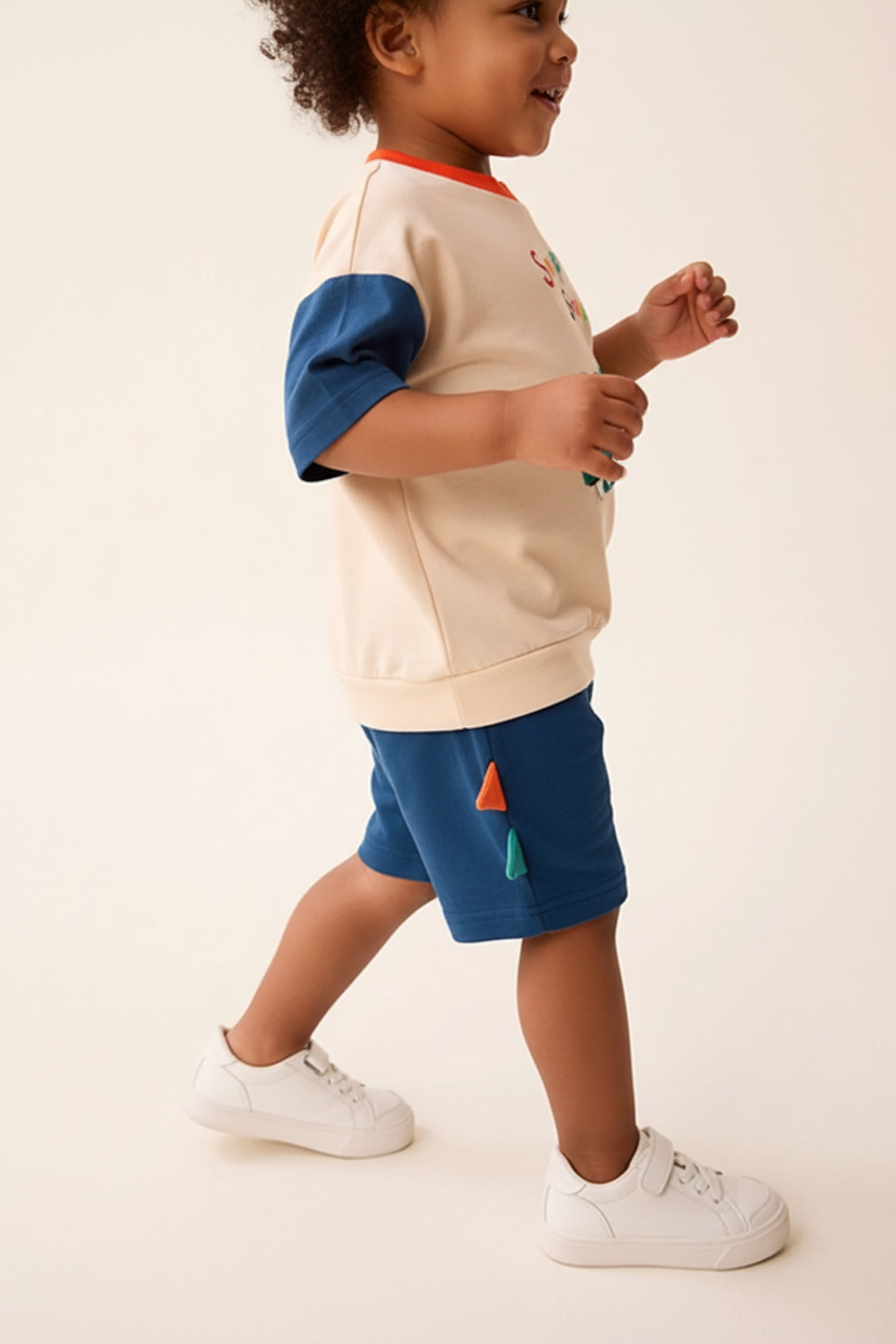 "Enfant 3 ans en pose active dynamique - Ensemble 2 pièces dinosaure sweat crème beige avec appliqué dinosaure vert adorable, texte 'Snap Snap' coloré multicolore, manches courtes bleu marine contrastantes, col rond orange - Short bleu marine avec pointes dinosaure colorées - Vue de profil latérale montrant silhouette complète de l'ensemble - Coupe confortable et ludique - Mouvement naturel et joueur - Fond blanc studio professionnel - Photo produit lifestyle premium BabyCoton Suisse - Style casual aventuri