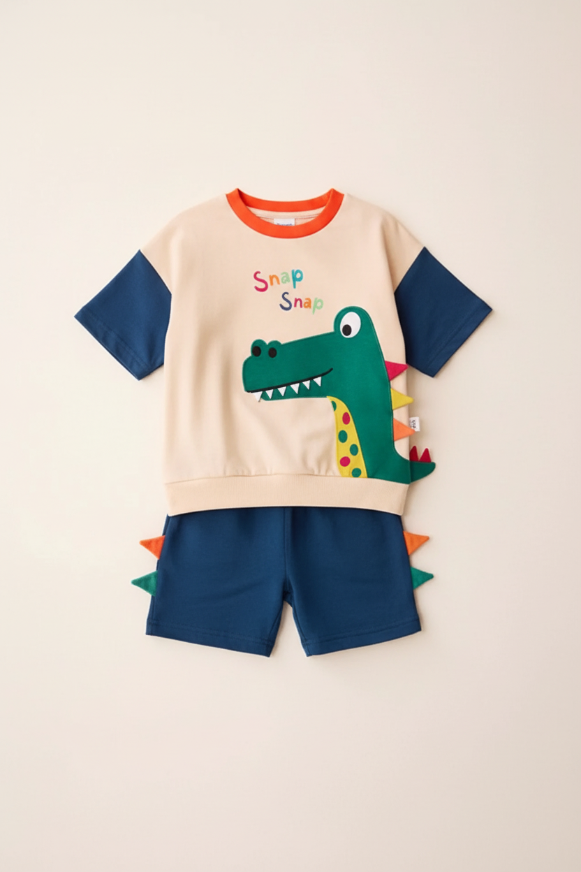"Ensemble 2 pièces dinosaure enfant - Vue de face complète à plat - Sweat beige crème avec texte 'Snap Snap' en lettres colorées (rose, bleu, vert), appliqué dinosaure vert avec dents blanches et détails multicolores, manches courtes bleu marine, col rond orange vif - Short bleu marine assorti avec pointes dinosaure colorées (orange, vert, rouge) au dos - Ensemble complet coordonné - Fond blanc professionnel - Photo produit e-commerce BabyCoton"