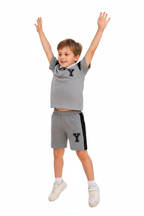 "Enfant 6 ans sautant de joie bras levés vers le haut - Ensemble polo et short gris chiné sportif - Polo col blanc avec bordures noires, logo Y noir appliqué sur poitrine, manches courtes avec détails noirs - Short coordonné gris avec bandes latérales noires contrastantes et logo Y - Vue trois-quarts angle diagonal montrant mouvement dynamique - Tenue athlétique confortable - Fond blanc studio professionnel - Photo produit lifestyle action BabyCoton Suisse - Style sportif décontracté garçon 3-8 ans"